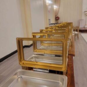 Gold-accented stainless steel chafing dishes elegantly arranged on a banquet table, set against a refined backdrop with floral decor, soft lighting, and neutral-toned drapery—perfect for upscale weddings, professional corporate receptions, and stylish brunch events in Houston.