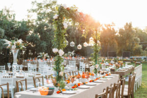 Outdoor wedding event with decorated banquet tables, floral arrangements, and hanging glass ornaments at sunset.