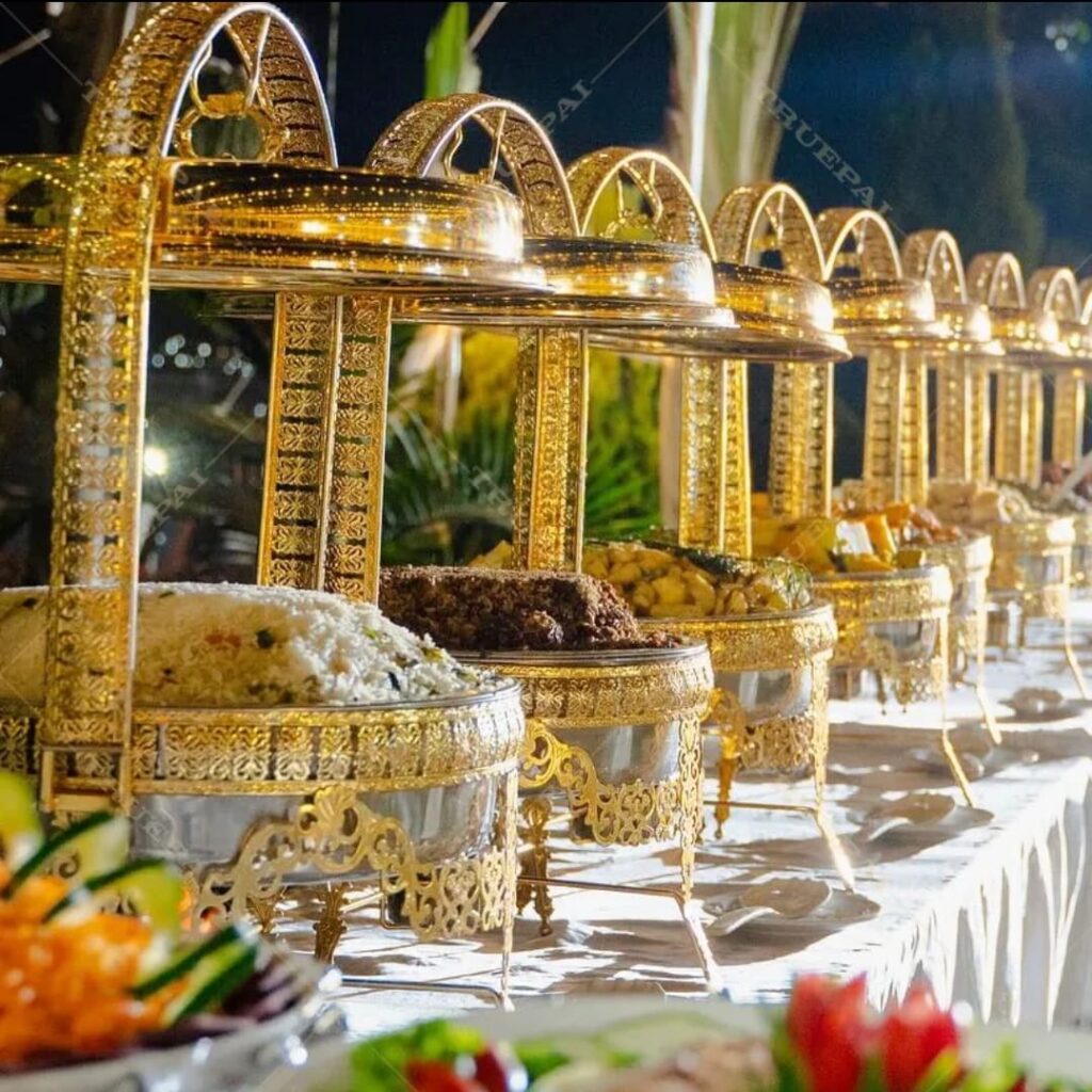 - Luxury Buffet Setup for Weddings & Banquets  Houston Party Service - Houston Party Rental Services Lavish outdoor buffet with ornate golden chafing dishes arranged on a white-clothed table, featuring a variety of dishes including rice, meats, and vegetables. The setup exudes luxury and warmth, ideal for upscale weddings, corporate banquets, or large nonprofit celebrations hosted by Houston Party Rental Services