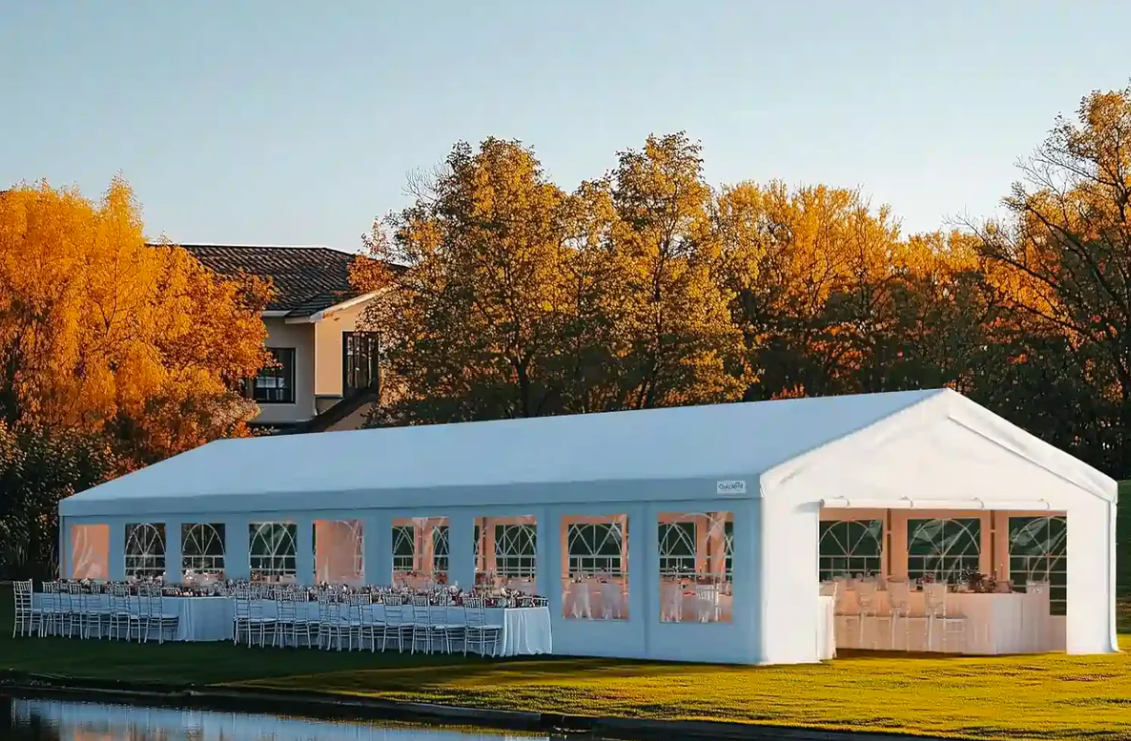 Large 20x70 white outdoor event tent with peaked roof and arched window panels, set up on grass near water. Interior features rows of tables and chairs for a formal gathering.